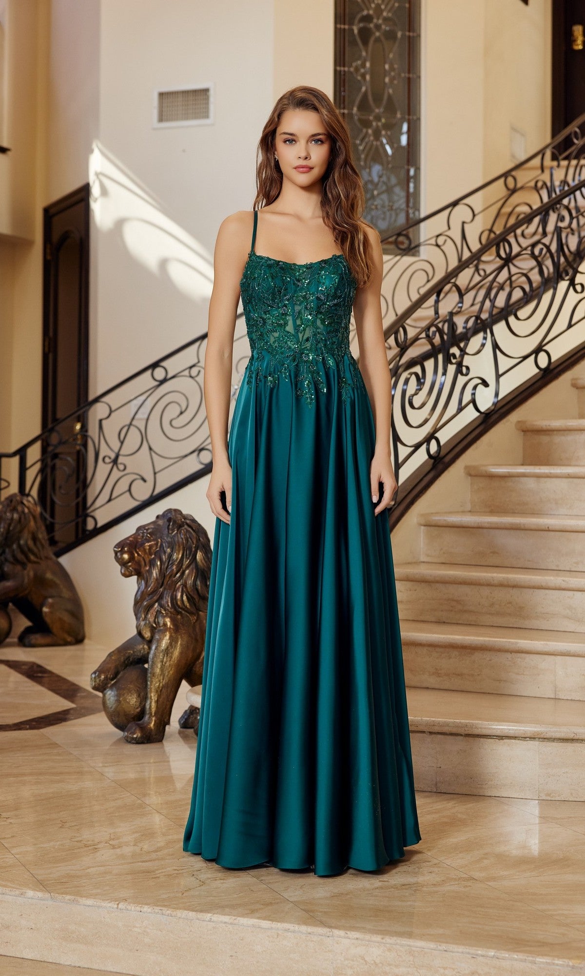 A woman wearing the Nox Anabel A1725 long prom dress, a sleeveless teal gown with sequin details, stands on a staircase with ornate railings and two lion statues in the background.